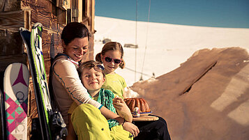 Mutter sitzt mit zwei Kindern an einer Skihütte und genießt die Sonne.