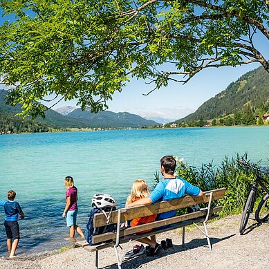 Familie macht eine Pause vom Fahrradfahren am See. Die Eltern sitzen auf einer Bank und die Kinder spielen am Wasser.